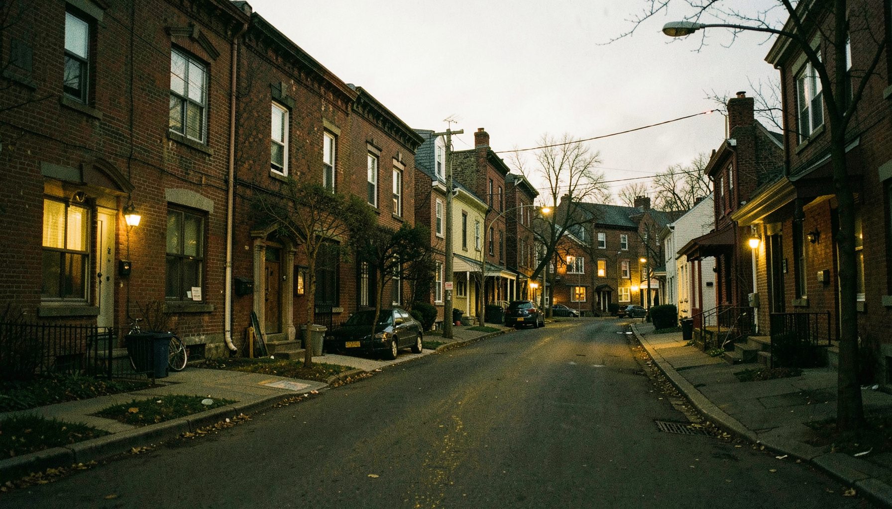 A dimly lit residential street in an older Toronto neighborhood at dusk, modest brick apartment buildings and small houses lining the road, warm yellow light spilling from a few windows, conveying quiet working-class life and economic struggle.